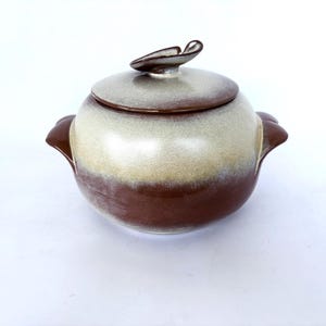 May include: A round ceramic lidded pot featuring a two-tone glaze with a dark brown base and a creamy off-white upper section. The pot includes two side handles and a lid with a curved, leaf-shaped handle on top, finished in a smooth glaze.