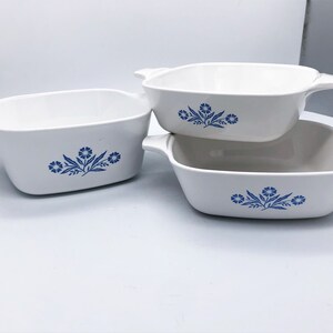 May include: Three white ceramic casserole dishes with blue floral designs. Each dish has a rectangular shape with rounded corners and handles on each side. The dishes are stacked on top of each other.