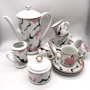 May include: A white porcelain coffee set with a pink, gray, and black abstract pattern. The set includes a coffee pot, creamer, sugar bowl, six cups, and saucers. Gold trim accents the edges of the pieces, creating an elegant look.