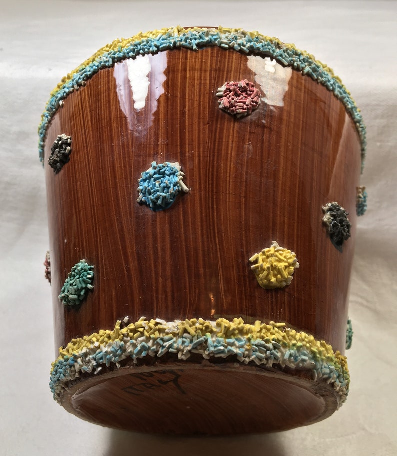 Puede incluir: Un recipiente de cer&aacute;mica marr&oacute;n con acabado brillante, decorado con confeti de colores alrededor de los bordes superior e inferior, y dispersos por la superficie. El confeti es en tonos azul, amarillo, rojo y negro. El recipiente tiene forma cil&iacute;ndrica.