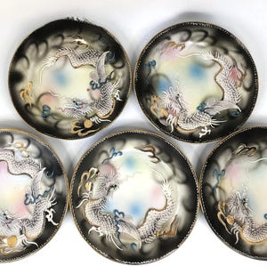 May include: Five decorative plates with a black and gold rim. Each plate features a central design of a white dragon with gold accents, set against a gradient background of black, white, and pastel colors. The plates are arranged in a circular pattern.