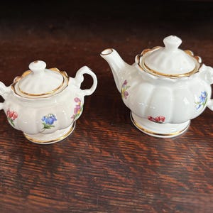 May include: A white porcelain teapot and a matching sugar bowl with lids. Both pieces feature a fluted body, gold-trimmed edges, and small painted floral motifs in red, blue, and purple. The teapot has a curved spout and a handle, while the sugar bowl has two side handles.