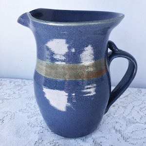 May include: A handmade ceramic pitcher in a speckled blue glaze. It features a wide spout, a curved handle, and a horizontal band of tan and gold. The pitcher is approximately 8 inches tall.
