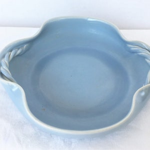 May include: A light blue ceramic bowl with a wavy rim and two braided rope handles. The bowl has a smooth, glossy finish and a shallow depth. The bowl is likely used for serving food or as a decorative piece.