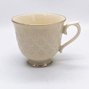 May include: Cream-colored teacup with a decorative embossed pattern and a gold rim. The handle has a unique, elegant design. The teacup sits on a small foot with a gold accent. A classic piece for tea or coffee.