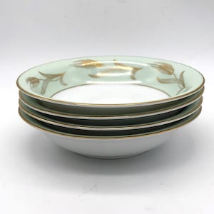 May include: A stack of four white and pale green bowls with gold trim and wheat stalk designs. The bowls have a shallow, rounded shape, and the gold accents add a touch of elegance to the tableware.