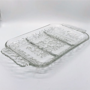 May include: A clear glass rectangular serving tray featuring three divided sections and integrated handles on both ends. The surface is decorated with an embossed floral pattern throughout the base and the raised edges of the dish.