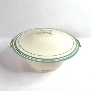 May include: Cream-colored serving dish with a lid. The dish has a rounded shape with a light green and black trim. It features a handle on the lid and two side handles. A classic piece for serving.
