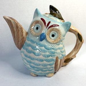 May include: A ceramic teapot shaped like an owl. The teapot is primarily light blue with brown and cream accents. The owl's eyes are large and round, with brown and red details. A brown branch forms the handle, and a green leaf sits on top.