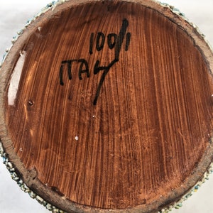 Puede incluir: Primer plano de la base de un jarr&oacute;n de cer&aacute;mica. La base es marr&oacute;n con textura de vetas de madera verticales. El texto "1001" e "ITALY" est&aacute; escrito con tinta negra. El borde est&aacute; decorado con peque&ntilde;as piedras de colores.