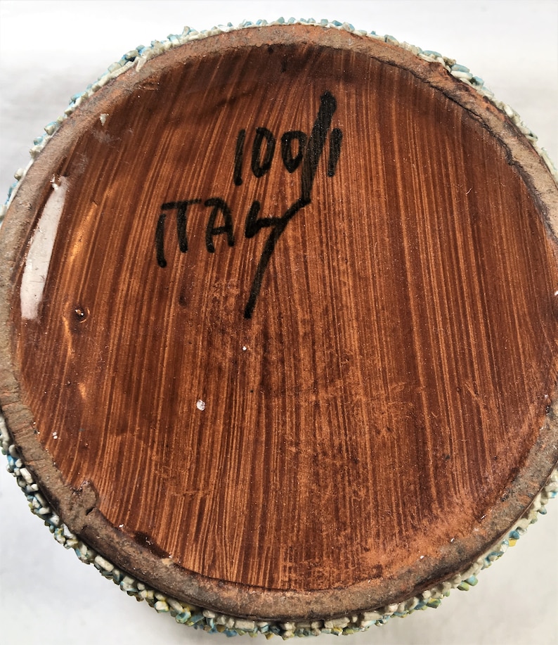 Puede incluir: Primer plano de la base de un jarr&oacute;n de cer&aacute;mica. La base es marr&oacute;n con textura de vetas de madera verticales. El texto "1001" e "ITALY" est&aacute; escrito con tinta negra. El borde est&aacute; decorado con peque&ntilde;as piedras de colores.