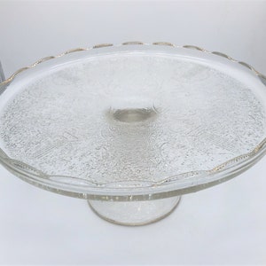 May include: Clear glass cake stand with a decorative etched pattern on the plate and a scalloped edge with gold trim. The stand has a short, pedestal base. A classic piece for displaying baked goods.