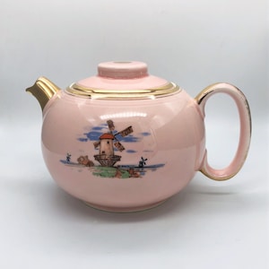 May include: A pink ceramic teapot with a windmill scene. The teapot has a gold-colored handle, spout, and trim. The windmill scene features a red-roofed windmill, blue sky, and green landscape.