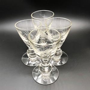 May include: Four clear glass cordial glasses with etched floral designs. Each glass has a round base, a stem, and a flared bowl. The glasses are arranged in a circular pattern against a dark background.