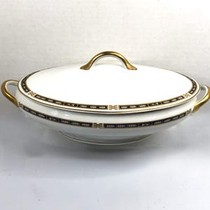 May include: White oval serving dish with a lid, featuring gold handles and trim. The dish has a decorative border with gold, navy blue, and black accents. A classic piece for serving food.
