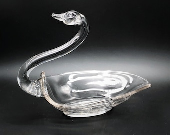 May include: A clear glass swan-shaped dish. The swan's neck curves gracefully, leading to a bowl-shaped body. The dish has a textured, rippled design on the base. The swan's head is detailed with a beak and eye, and the dish is set against a black background.