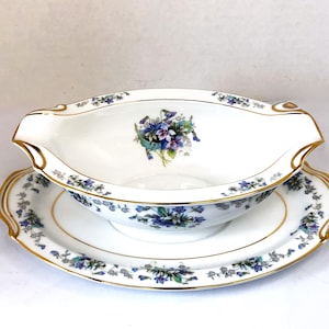 May include: White porcelain gravy boat and underplate set. The boat features a floral design with blue and purple flowers, gold trim, and a spout. The underplate mirrors the design. A classic addition to any dining table.