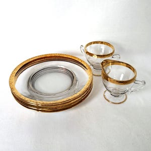 May include: A vintage glass tea set with gold trim. The set includes a stack of saucers, two teacups, and a creamer. The rims of the cups, saucers, and creamer are accented with a gold band. The set is displayed on a white surface.