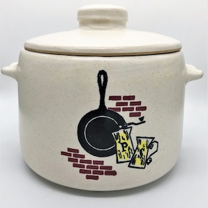 May include: Cream-colored ceramic cookie jar with a lid and side handles. The jar features a black skillet graphic, brick accents, and yellow and black text. A vintage kitchenware item.