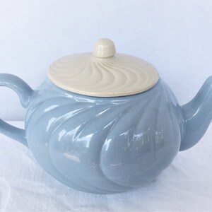 May include: A light blue ceramic teapot with a matching handle and spout. The lid is a cream color with a swirl design. The teapot is sitting on a white surface. A classic tea accessory.