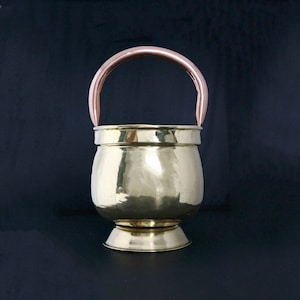 May include: A brass bucket with a copper handle. The bucket has a rounded body and a flared base. The handle is curved and has a smooth finish.