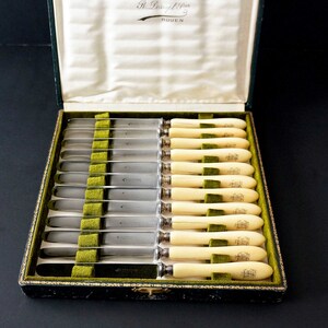 Puede incluir: Un juego de 12 cuchillos de mantequilla de plata con mangos de marfil en una caja forrada de terciopelo verde. La caja est&aacute; etiquetada "Monbenoult R. Dessupt Frere Rouen".