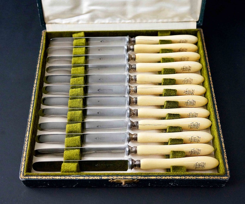Puede incluir: Un juego de 12 cuchillos de mantequilla de plata con mangos de marfil en una caja forrada de terciopelo verde. Los mangos est&aacute;n grabados con un escudo de armas.