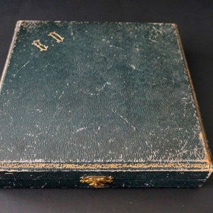 Puede incluir: Una caja rectangular de color verde oscuro con ribetes dorados y un cierre de lat&oacute;n. La caja tiene las letras "B.D." en relieve en la parte superior.
