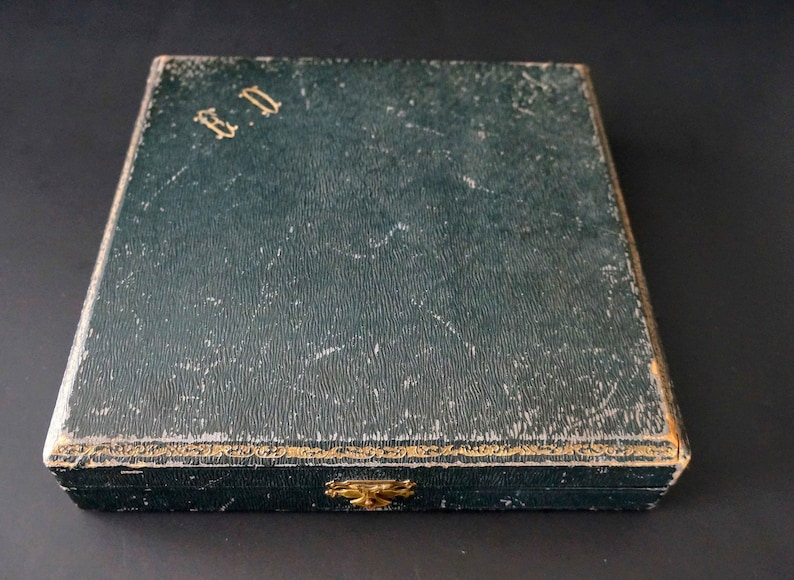 Puede incluir: Una caja rectangular de color verde oscuro con ribetes dorados y un cierre de lat&oacute;n. La caja tiene las letras "B.D." en relieve en la parte superior.