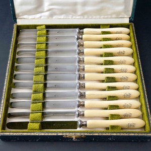 Puede incluir: Un juego de 12 cuchillos de mantequilla de plata con mangos de marfil en una caja forrada de terciopelo verde. Los mangos est&aacute;n grabados con un monograma.