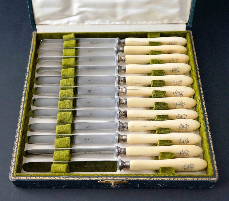 Puede incluir: Un juego de 12 cuchillos de mantequilla de plata con mangos de marfil en una caja forrada de terciopelo verde. Los mangos est&aacute;n grabados con un monograma.