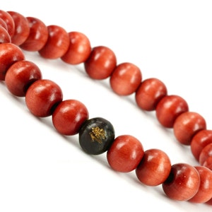 May include: A red wooden beaded bracelet with a black bead featuring gold lettering. The bracelet is made of round beads and is approximately 8 inches in length.