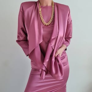 May include: A mauve satin dress with a matching satin jacket. The jacket is draped over the shoulders and tied at the waist. The dress has a fitted silhouette and a scoop neckline. The model is wearing a gold chain necklace.