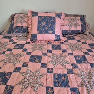 May include: A quilted bedspread and pillow set with a star pattern. The quilt and pillows have a pink background with navy blue squares and floral star designs. The pillows have frilled edges.