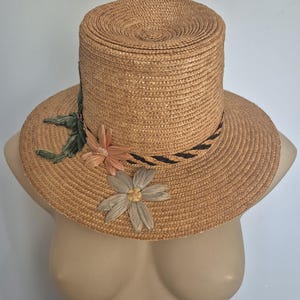 May include: A brown straw hat with a wide brim and a tall crown. The hat is decorated with woven flowers in shades of green, pink, and white.