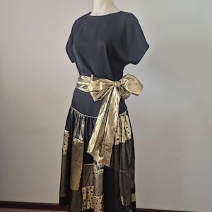 May include: Black dress with a gold metallic bow belt. The skirt has a patchwork design with gold and black fabrics. The dress features short sleeves and a round neckline. The dress is displayed on a mannequin.