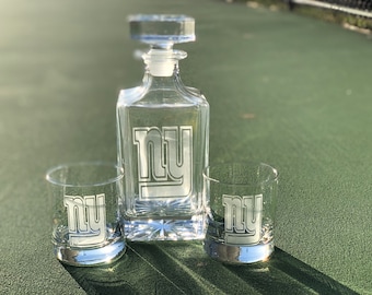 May include: A clear glass decanter and two matching glasses with an etched "NY" logo. The glasses and decanter are on a green surface.