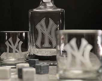 May include: A set of three clear glass decanters with a white etched New York Yankees logo. The decanters are on a gray surface with gray and white stones.