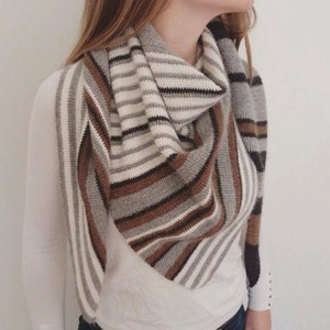 Puede incluir: Una bufanda de crochet a rayas en tonos de marrón, gris, blanco y negro. La bufanda está doblada en forma triangular y drapeada alrededor del cuello de una persona.