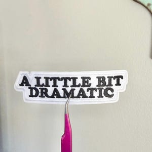 May include: A white patch with black embroidered text that reads "A LITTLE BIT DRAMATIC." The patch is held by pink tweezers. The patch has a white border and is rectangular with curved edges.