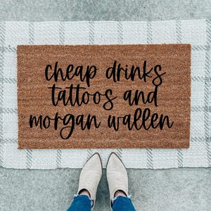 May include: A brown doormat with the text "cheap drinks tattoos and morgan wallen" in black lettering.