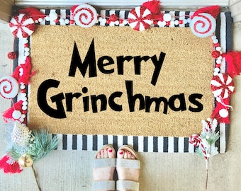 May include: A doormat with the words "Merry Grinchmas" in black. The mat is bordered with a black and white striped pattern. It is decorated with red and white candy cane and peppermint accents. The feet of a person wearing silver sandals are visible.
