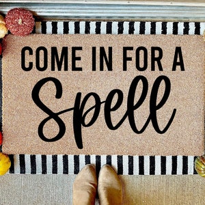 May include: A brown coir doormat with black and white striped edging. The doormat has the text "Come in for a Spell" in black lettering.