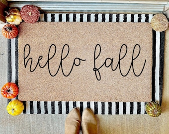 May include: A brown coir doormat with black and white stripes around the edges. The doormat has the text "hello fall" written in black cursive font.