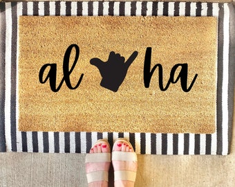 May include: A brown coir doormat with black text that reads "Aloha" with a black hand gesture in the middle.