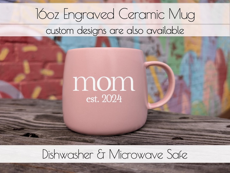 Op de afbeelding: Een roze keramische mok van 473 ml met het woord "mom" en "est. 2024" in witte letters. De mok heeft een gebogen vorm en een oor. Tekst bovenaan luidt "16oz Gegraveerde Keramische Mok" en "aangepaste ontwerpen zijn ook beschikbaar."