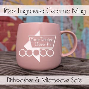 Op de afbeelding: Een roze 473ml gegraveerde keramische mok met een handvat. De mok heeft een wit ontwerp met de tekst "Your Design Here" en is gemarkeerd met "Dishwasher & Microwave Safe". De mok staat tegen een kleurrijke achtergrond.