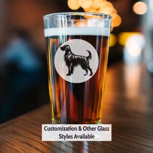 May include: A glass of beer with a black silhouette of a dog on a white circle sticker. The text "Customization & Other Glass Styles Available" is below the glass.