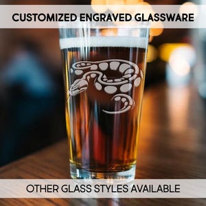 May include: A pint glass filled with a dark beverage, possibly beer, featuring a white engraved snake design. The glass has the text "CUSTOMIZED ENGRAVED GLASSWARE" at the top and "OTHER GLASS STYLES AVAILABLE" at the bottom.