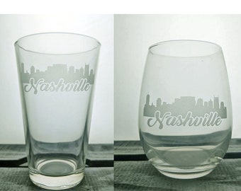 May include: Two clear glass tumblers with the word "Nashville" and a skyline etched on the front. One is a pint glass, the other a stemless wine glass. Both are empty.